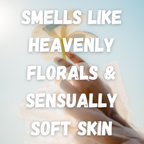A woman's hand emerging from a sheer white sleeve holds a white flower with the sunny blue sky in the background. The words "smells like heavenly florals & sensually soft skin" overlay the image.